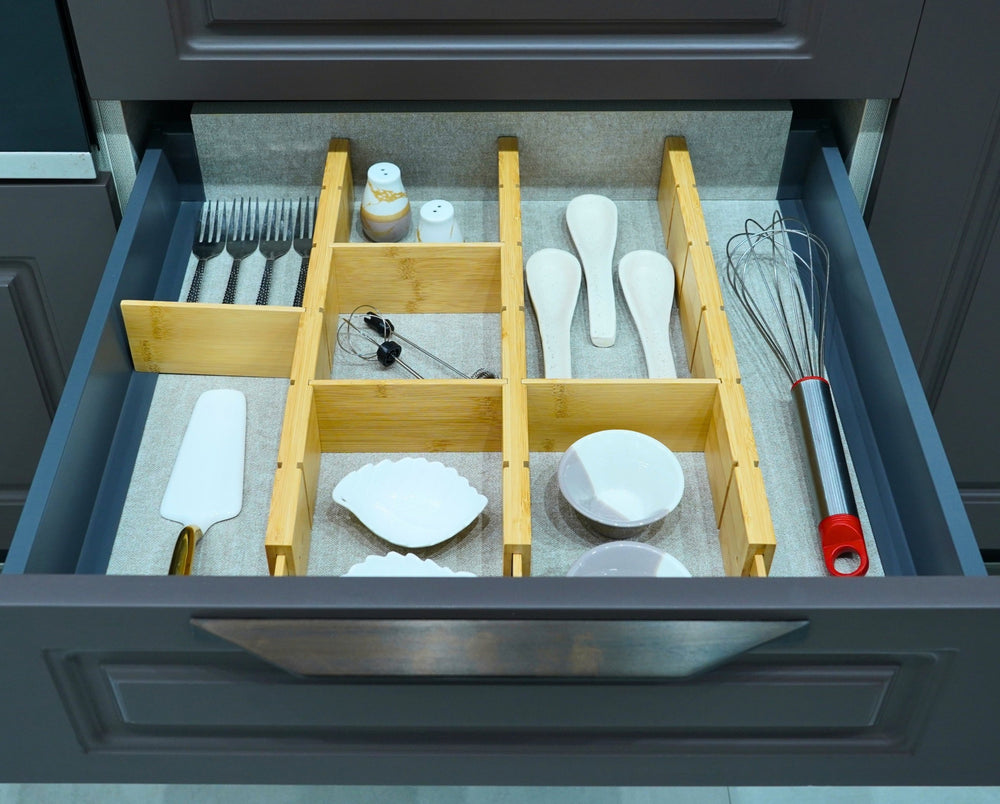 Expandable bamboo drawer dividers organising utensils, a whisk and small dishes in a grey kitchen drawer